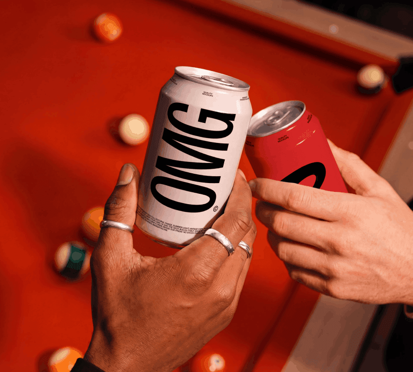 Two hands clinking soda can mockups over a red pool table, a white can with bold OMG type meeting a red can, billiard balls scattered behind.