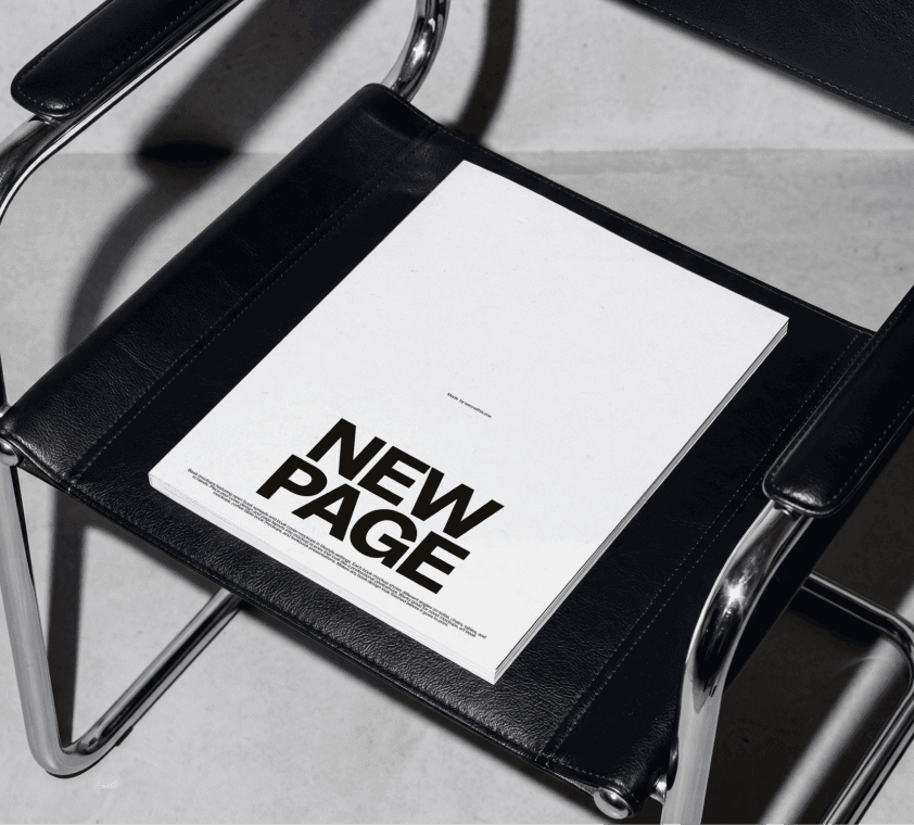 Free softcover book mockup resting on a black leather Bauhaus-style chair with chrome frame and concrete floor.