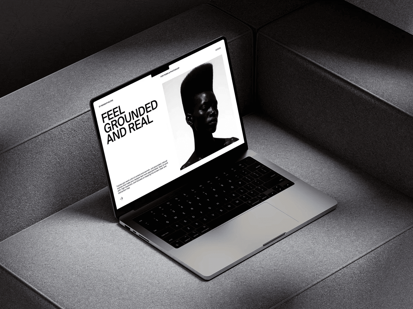 Modern MacBook laptop mockup on grey fabric steps displaying a minimalist website with bold typography and a black-and-white portrait