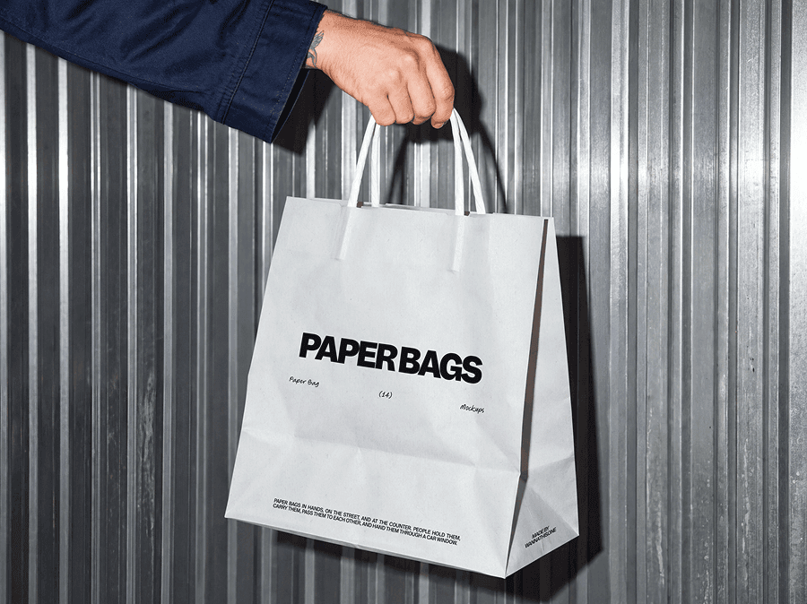 Paper shopping bag mockup hanging from one hand in front of a ribbed metal wall