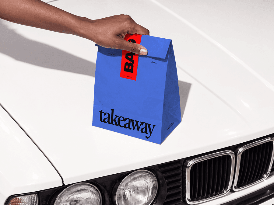 Folded takeaway paper bag mockup steadied by one hand on the hood of an old car