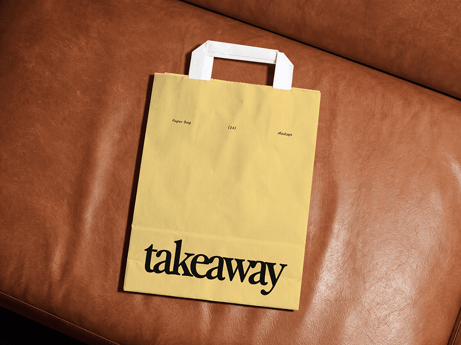 Paper shopping bag mockup with handles sitting upright on a brown leather sofa