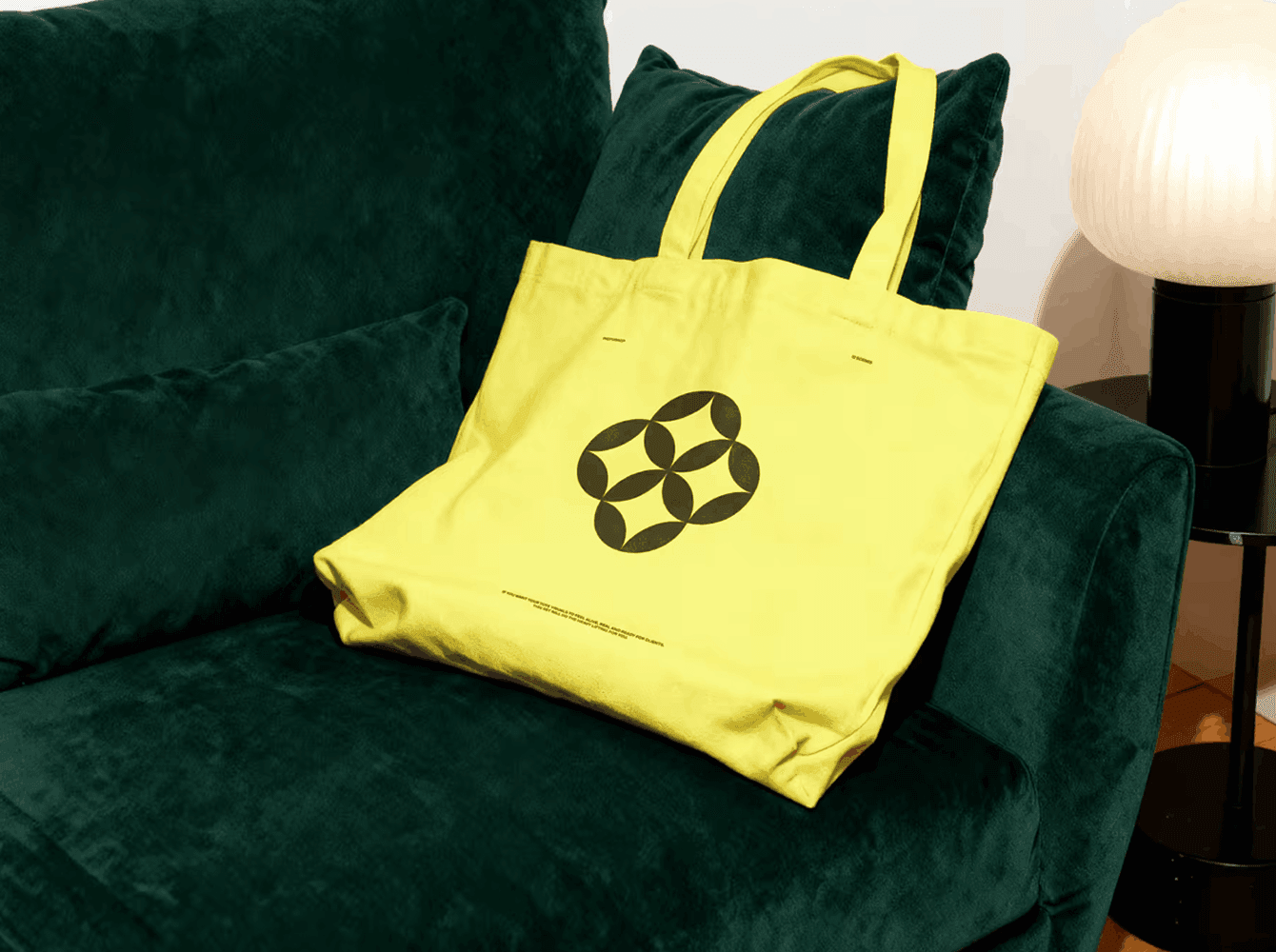Yellow canvas tote bag mockup with geometric print, resting on a dark green velvet sofa beside a warm lamp — a lifestyle scene for presenting brand identity on merch.